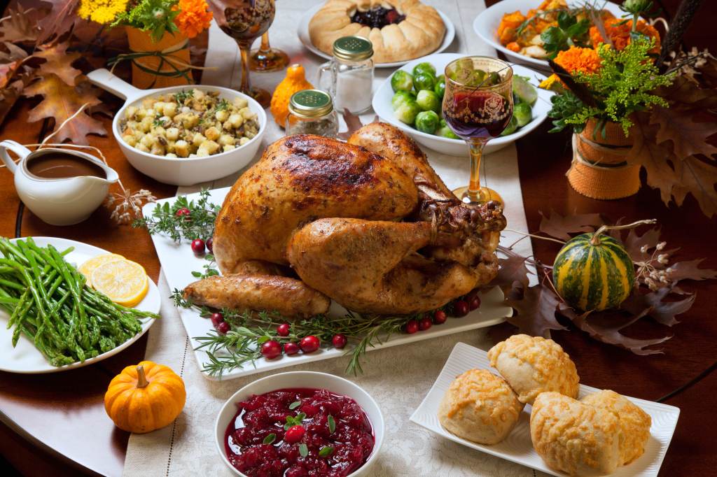 Roast turkey garnished with cranberries on a rustic Thanksgiving table decorated with pumpkins, gourds, asparagus, brussels sprouts, roasted vegetables, pie, flowers and candles.